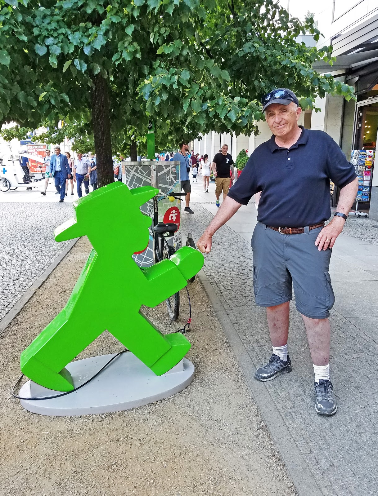 Outdoor advertising figure of Ampelmann. 1 Mężczyzna stojący obok dużej zielonej figury przedstawiającej chodzącego człowieka na chodniku, w tle drzewa i ludzie.