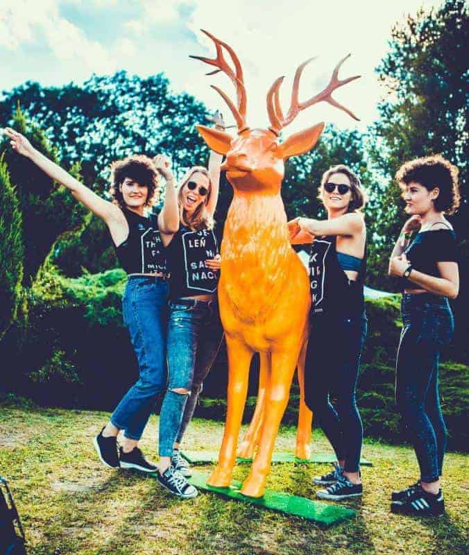 Cztery kobiety pozują przy pomarańczowej figurze jelenia na tle zieleni.