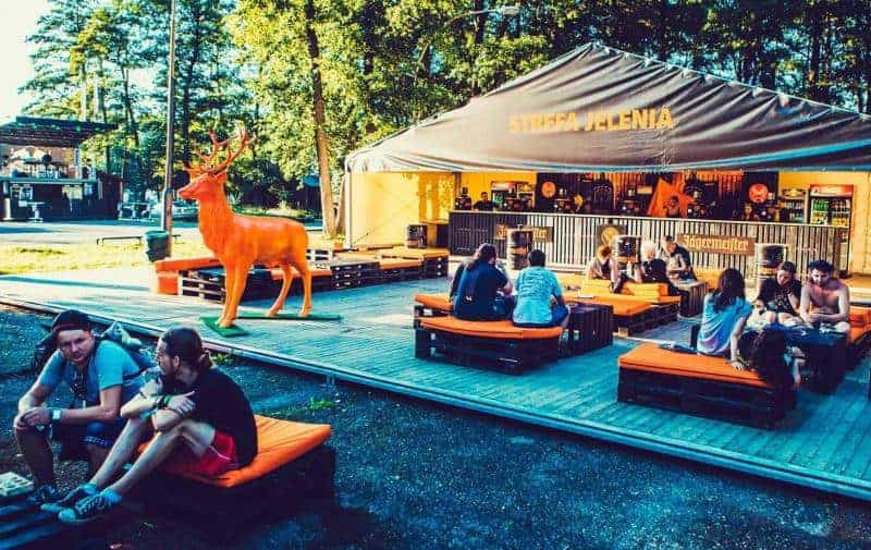 Ludzie siedzący na pomarańczowych poduszkach na drewnianej platformie przed namiotem z napisem 