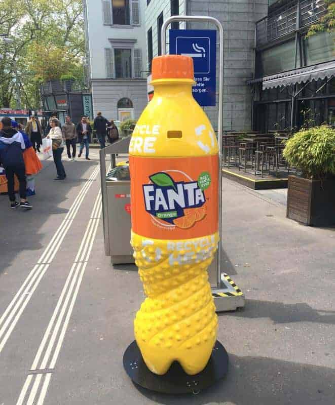 Giant Fanta bottle-shaped recycling bin placed on a city street with pedestrians and buildings in the background.