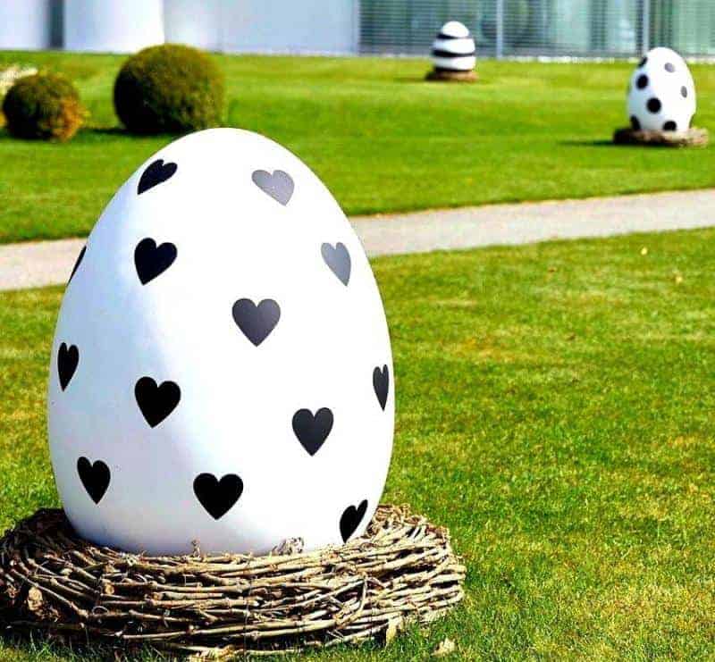 oryg_064490685_1129949550544944_1465385417682452480_o Giant white egg decorated with black heart shapes, positioned on a grassy area with several similar eggs in the background.