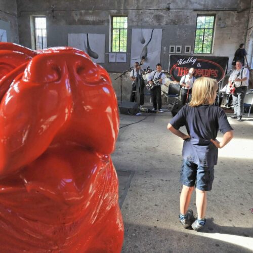 Children watch a band perform inside a rustic building with a large red sculpture in the foreground.