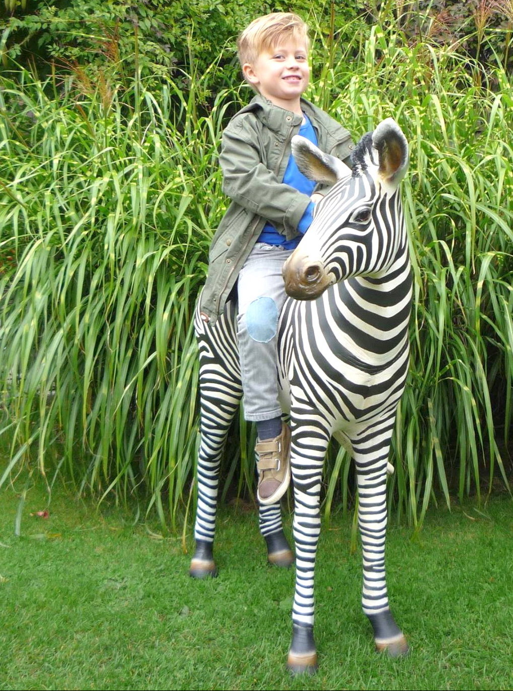 Zebra realistic life size fiberglass figure 2 A child sits on a zebra sculpture in front of tall grass.