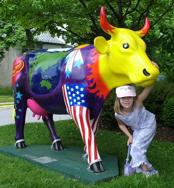 Fiberglass Cow Life Size Statue - Single Color 12 A girl posing next to a brightly colored cow statue with a world map, American flag, and assorted vibrant designs painted on it.