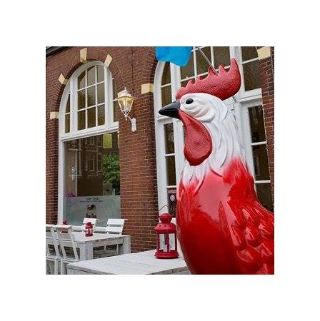 Large red and white statue of a rooster outside a building with arched windows and outdoor seating.