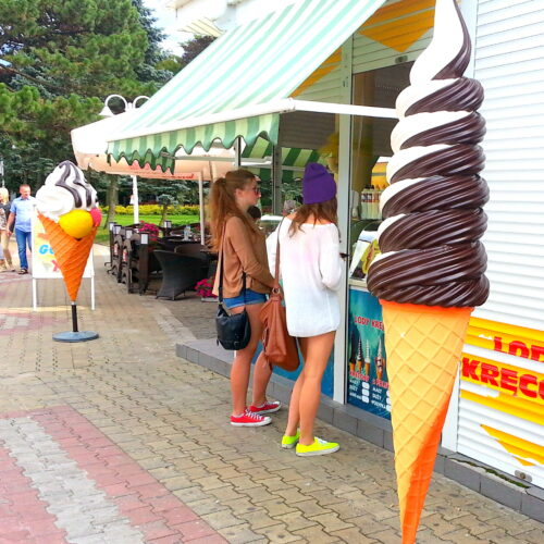 Two people stand in line at an ice cream stand with large ice cream cone decorations, one chocolate vanilla swirl and the other with scoops of various flavors.