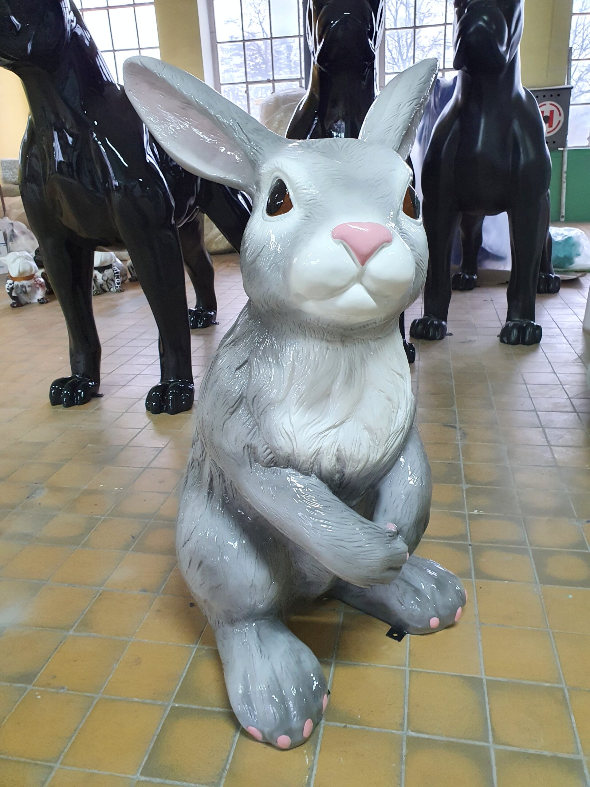 Large Rabbit Fiberglass Statue - Single Color 4 Large statue of a gray rabbit with pink nose and paws, standing on its hind legs, surrounded by black statues of dogs in an indoor setting.