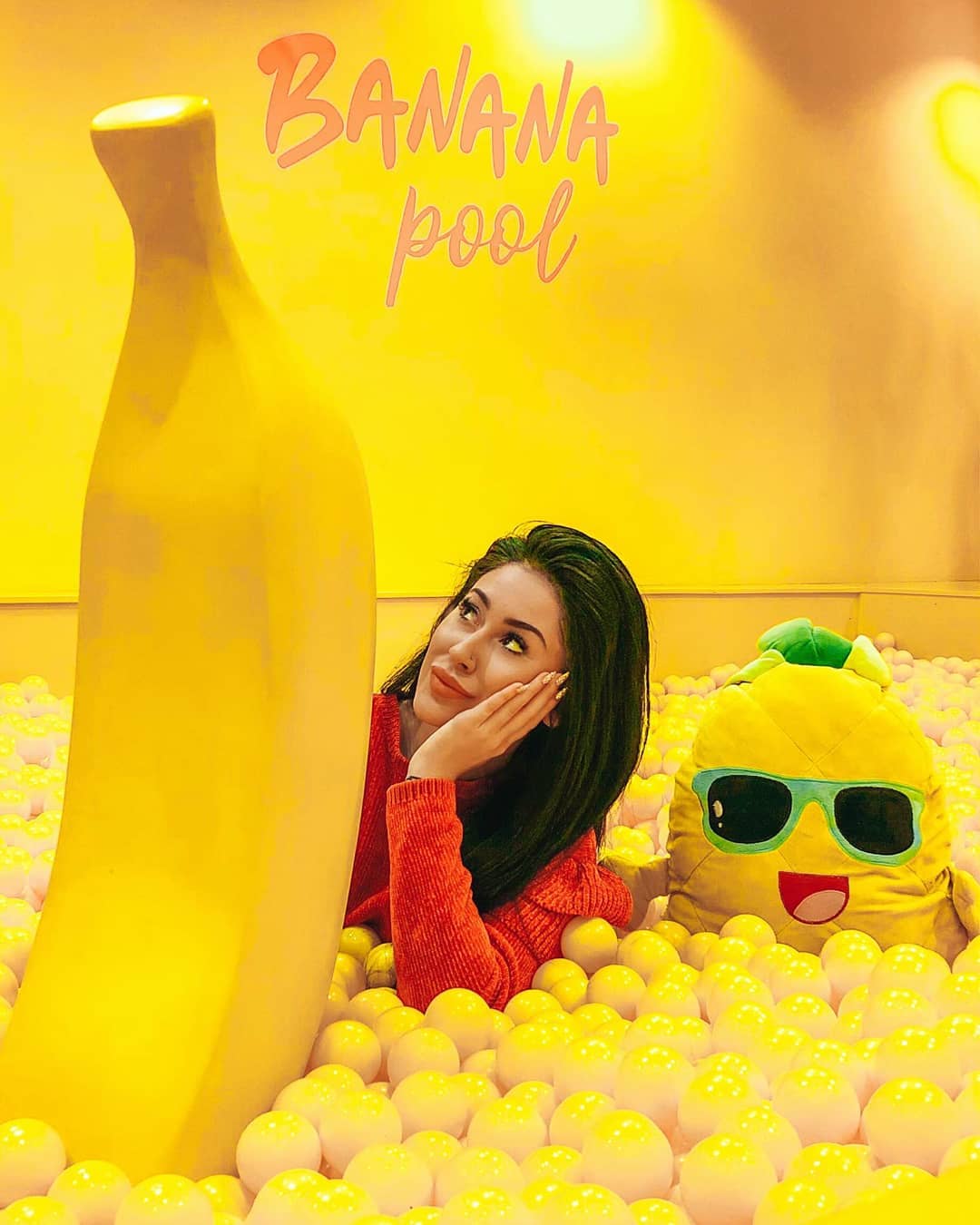 Realistic Large Banana Fiberglass Model 3 Woman lying in a yellow ball pit next to a large banana sculpture and a smiling stuffed toy wearing sunglasses, with