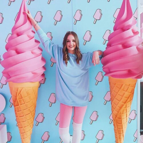 Woman posing between two oversized pink ice cream cones with a blue and pink popsicle-themed background.