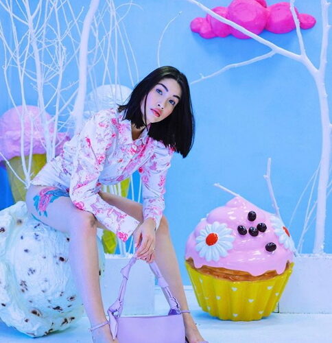 A woman with shoulder-length dark hair poses in a colorful, whimsical setting featuring oversized cupcake and ice cream decorations, wearing a floral top and shorts, holding a lilac handbag.