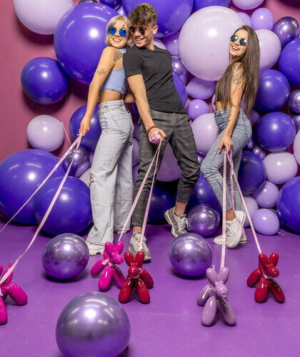 Three people pose with balloon dogs on leashes in a room filled with large purple and white balloons.