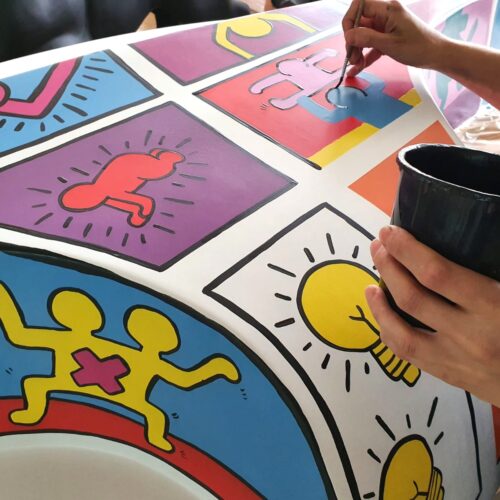 A person painting a colorful mural featuring cartoon-like figures and symbols, including yellow light bulbs, and various human shapes in different colors and poses.