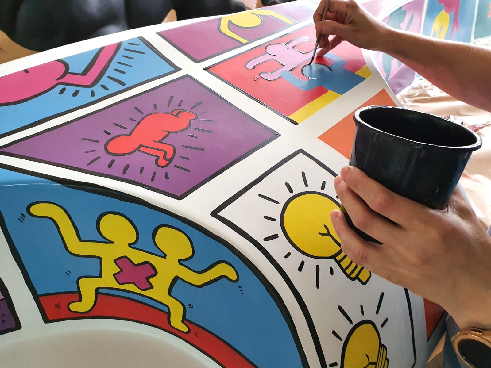 Shopping center fiberglass bench 2 A person painting a colorful mural featuring cartoon-like figures and symbols, including yellow light bulbs, and various human shapes in different colors and poses.