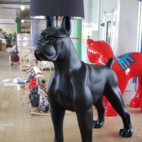 Black dog sculpture with a black lampshade on its head, standing on a tile floor in a workshop.