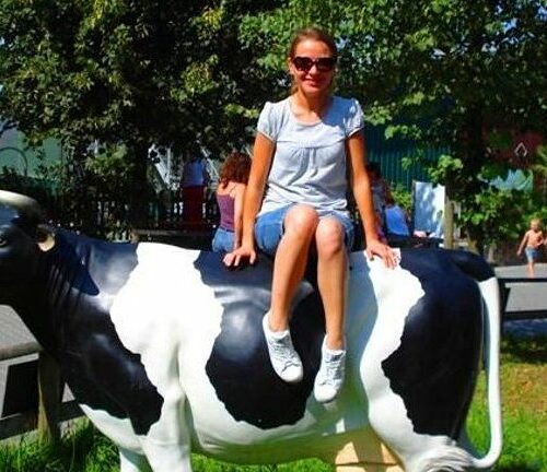 Person sitting on a black and white cow sculpture outdoors in a sunny park.
