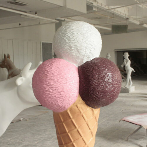 Large sculpture of a three-scoop ice cream cone with vanilla, strawberry, and chocolate scoops in an indoor workshop setting.