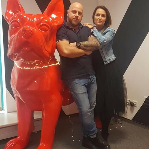 Man and woman posing with large red dog statue in a modern indoor setting.