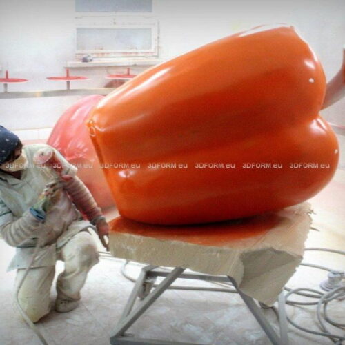 Person spray-painting a large orange bell pepper model in a workshop.