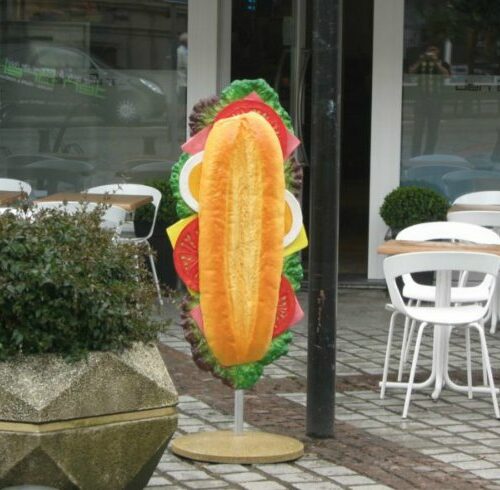 Large sandwich model on a pole in front of an outdoor café with white chairs and tables.
