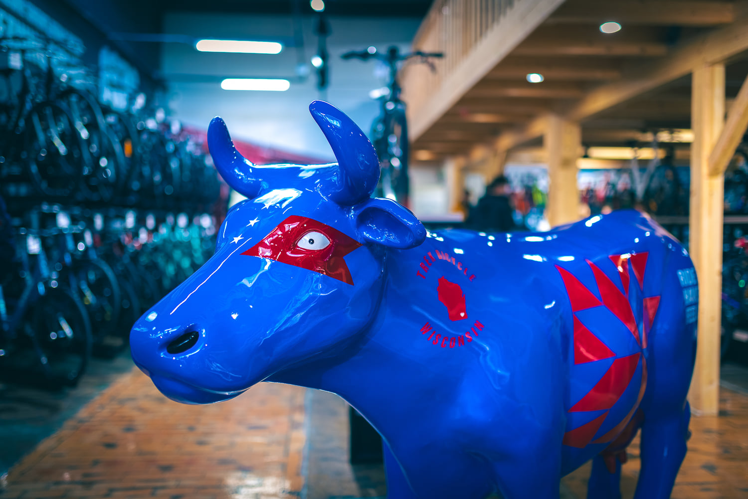 Fiberglass Cow Life Size Statue - Single Color 2 Blue cow sculpture with red painted designs on its eyes and body, placed inside a bicycle store with wooden flooring and racks of bicycles in the background.