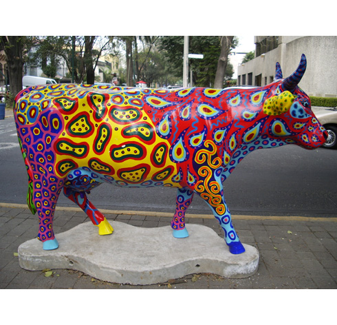 Fiberglass Cow Life Size Statue - Single Color 14 Colorful art installation of a cow with vibrant yellow, red, blue, and orange patterns on its body, standing on a concrete platform by a roadside.