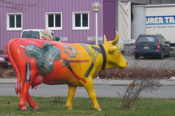 Fiberglass Cow Life Size Statue - Single Color 15 Colorful cow statue painted with various tools on its body, placed on a grassy patch near a road with vehicles and a purple building in the background.