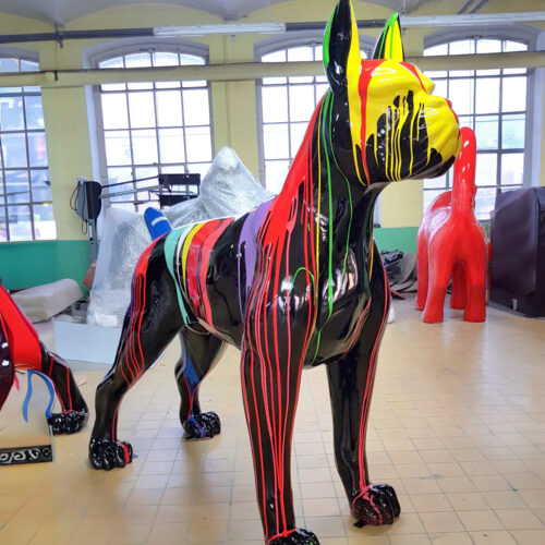 Large black dog sculpture with rainbow-colored paint drips in a room with large windows and other colorful animal sculptures in the background.