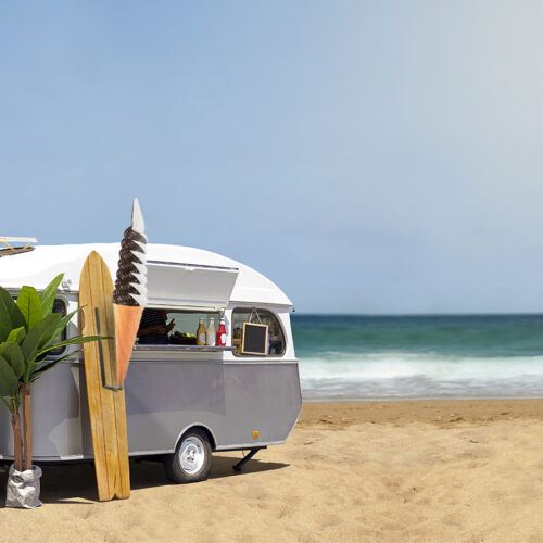 Food truck on a sandy beach with a surfboard and tropical plant nearby, ocean waves in the background.
