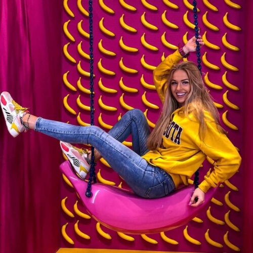 Woman sitting on a pink swing shaped like a banana, smiling, wearing a yellow hoodie and jeans, with a background of yellow bananas on a pink wall.