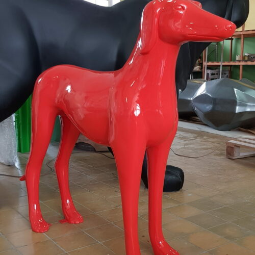A bright red, glossy statue of a dog with elongated legs and a streamlined body standing on a tiled floor, with parts of other large dog statues visible in the background.