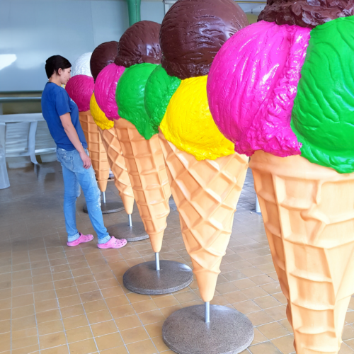 Person standing next to large ice cream cone sculptures with colorful scoops in a tiled indoor space.