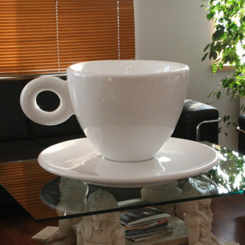 A giant, white teacup and saucer sculpture placed on a modern glass coffee table with pharaoh figurine supports in a living room.