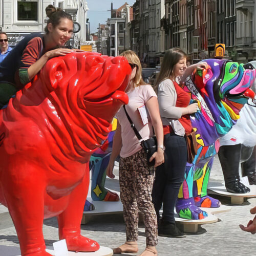 People interacting with large colorful dog statues on a street with buildings in the background.