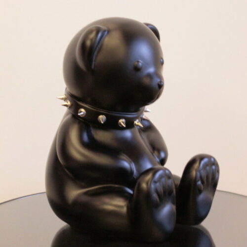 Black teddy bear figurine with a spiked collar around its neck, sitting on a black reflective surface.
