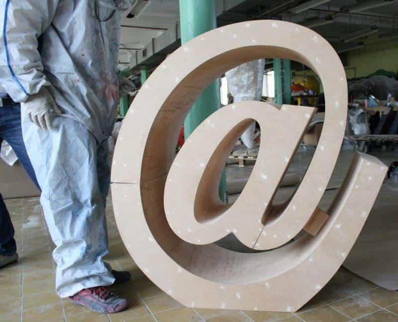 Email writing sign 2 Duża rzeźba symbolu małpy (@) w warsztacie, obok której stoi osoba w kombinezonie ochronnym.