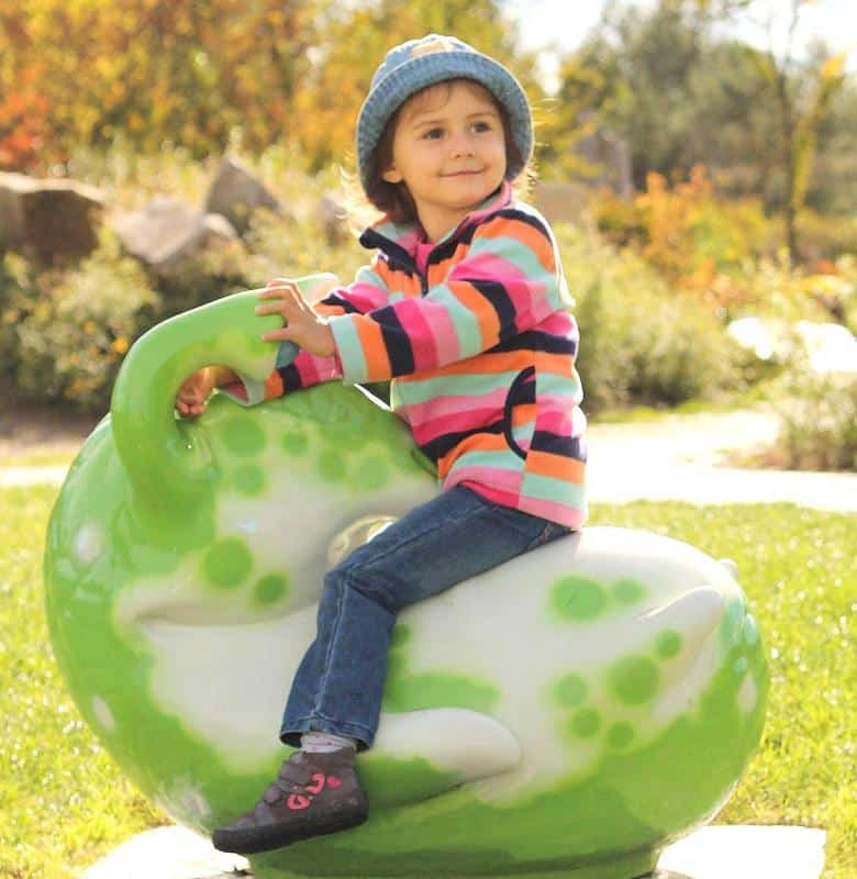 Dziecko w kolorowej bluzie i niebieskiej czapce siedzi na zielonej huśtawce w kształcie dinozaura na placu zabaw.