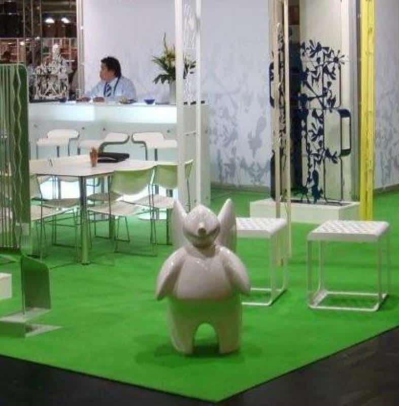 Exhibition booth with green carpet, white furniture, decorative plants, and a small statue of a white creature with wings in the foreground.