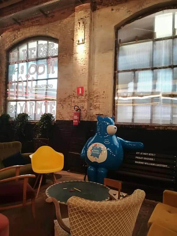 oryg_064490685_1129949550544944_1465385417682452480_o Interior of a cozy room with colorful chairs, a round table, exposed brick walls, and large windows. A large blue bear statue is sitting on a bench.