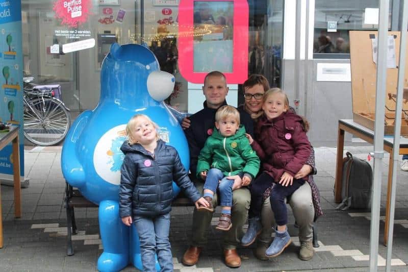 Bear on a Bench 2 Rodzina z trójką dzieci pozuje do zdjęcia obok dużej niebieskiej maskotki w przestrzeni publicznej.