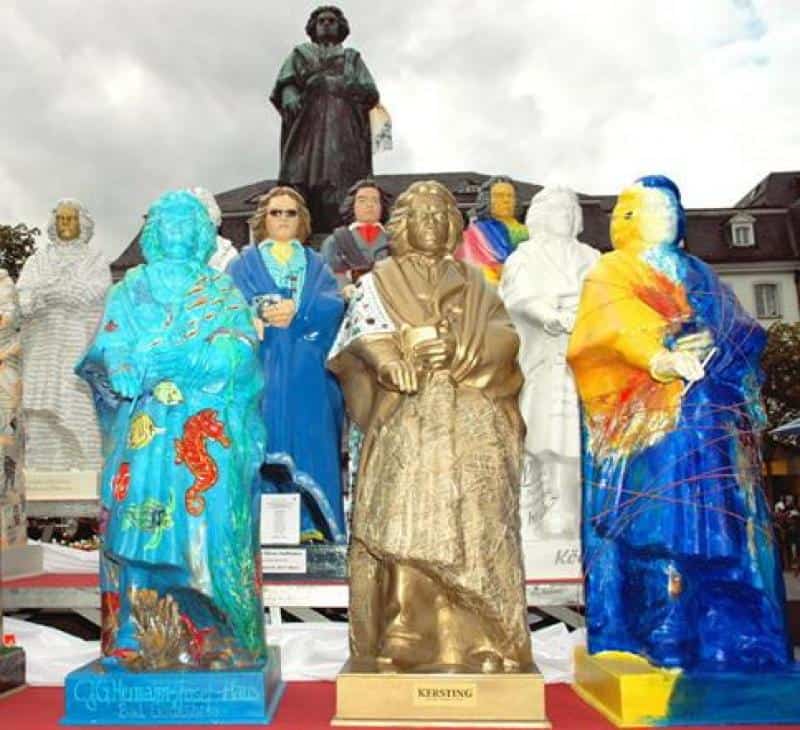 oryg_064490685_1129949550544944_1465385417682452480_o Colorful statues of cloaked figures displayed outdoors, with a larger dark statue in the background.