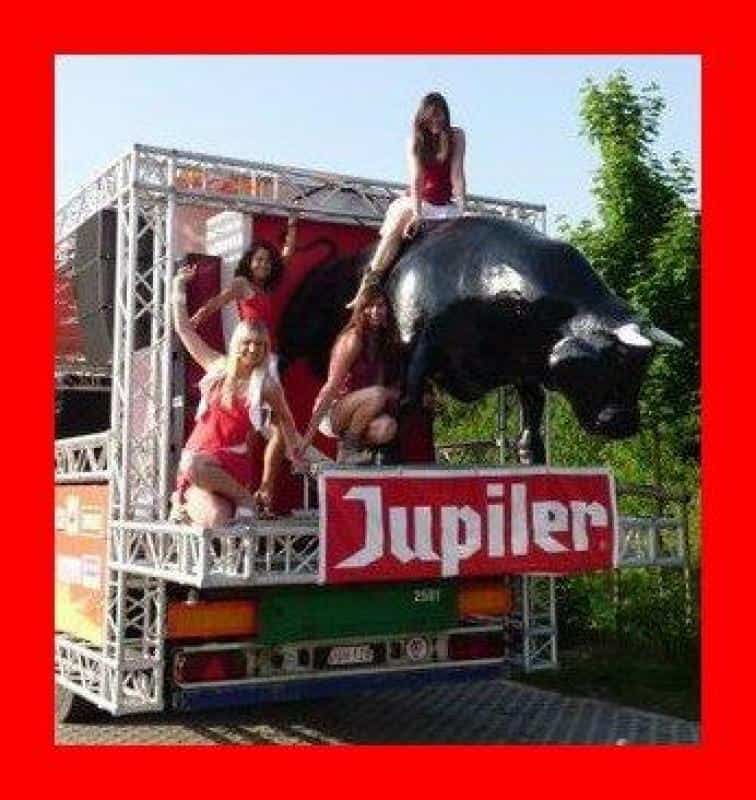 oryg_064490685_1129949550544944_1465385417682452480_o Four women in red outfits posing on a Jupiler beer truck adorned with a black bull statue.