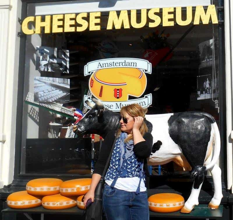 oryg_064490685_1129949550544944_1465385417682452480_o Woman standing beside a cow statue outside the Amsterdam Cheese Museum with wheels of cheese displayed.
