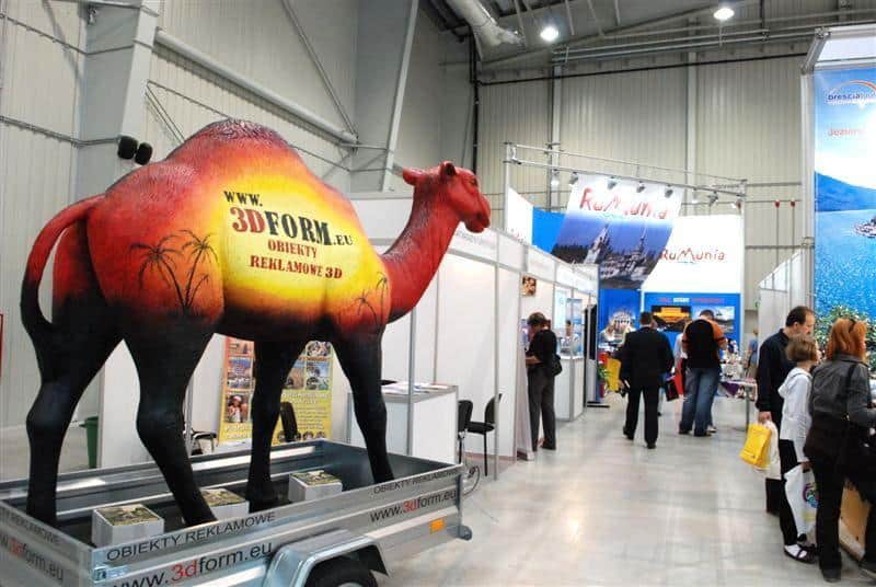 Mobile camel mock-up 4 Stoisko wystawowe z dużą figurą wielbłąda promującą obiekty reklamowe 3D; w tle kilka stoisk z banerami i ludzie oglądający ekspozycje.