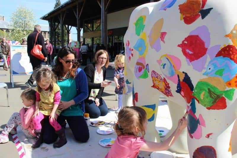 oryg_064490685_1129949550544944_1465385417682452480_o People, including children, painting a large, white elephant statue with colorful designs outdoors.
