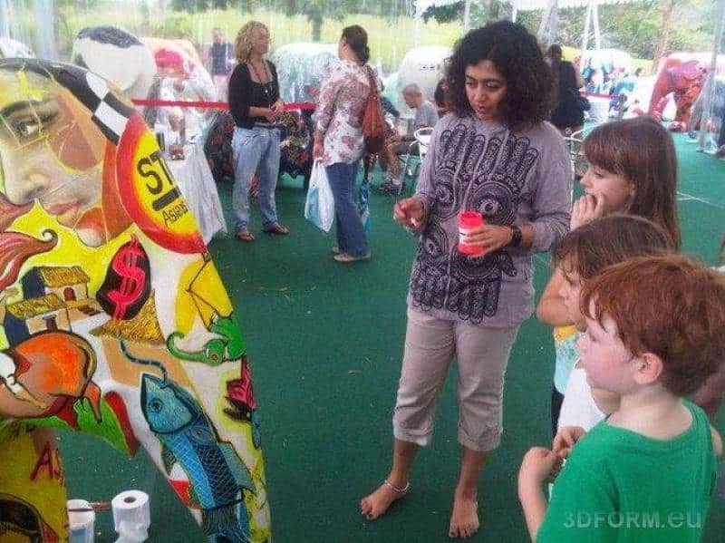 Large figures painted by children 2 Kobieta i dzieci stojące przed kolorowym muralem na wystawie plenerowej.