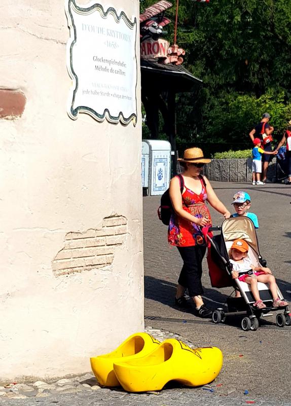 Kobieta z wózkiem dziecięcym przechodzi obok dużych żółtych drewniaków przy ścianie z francuskim napisem.