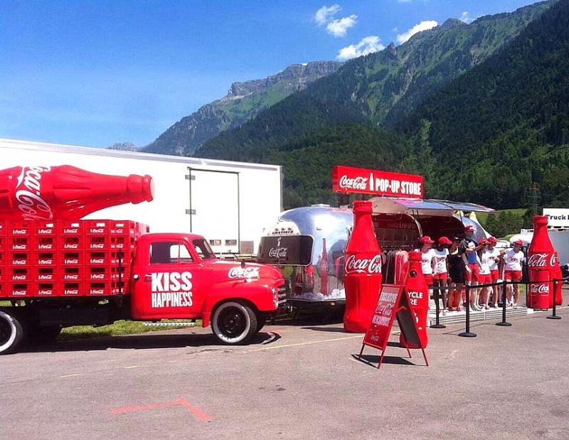 Coca-Cola advertising truck 3 Czerwony samochód Coca-Coli z dużą butelką na przyczepie i sklepem pop-up w tle, stojący na parkingu w otoczeniu gór.