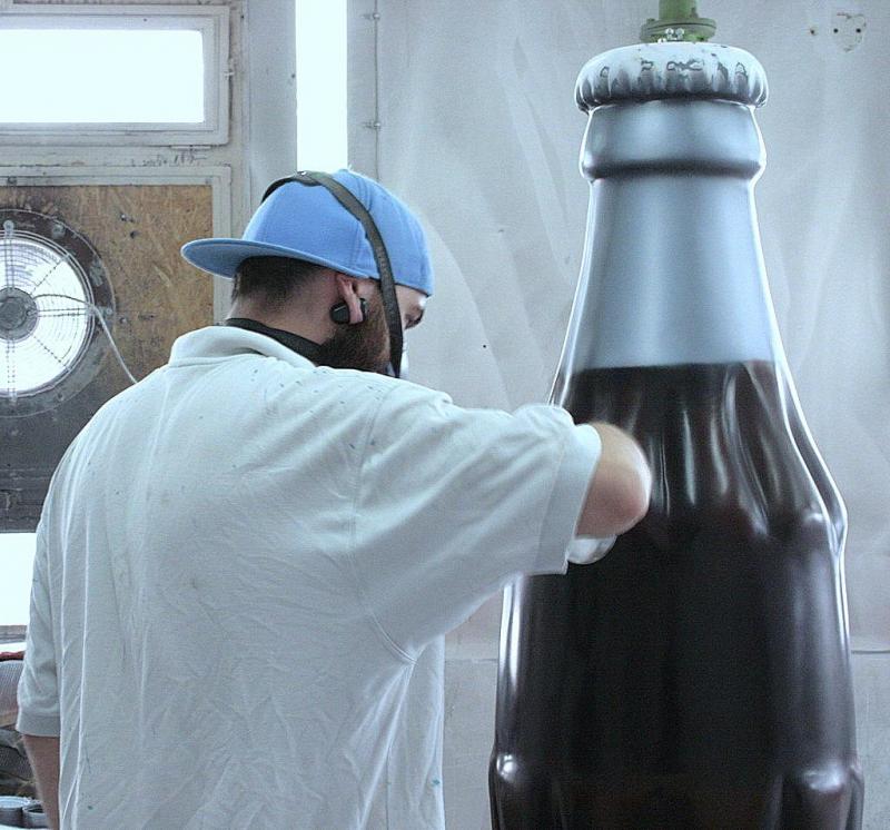Hanging Coca-Cola bottles. 3 Mężczyzna w niebieskiej czapce lakieruje dużą butelkę.