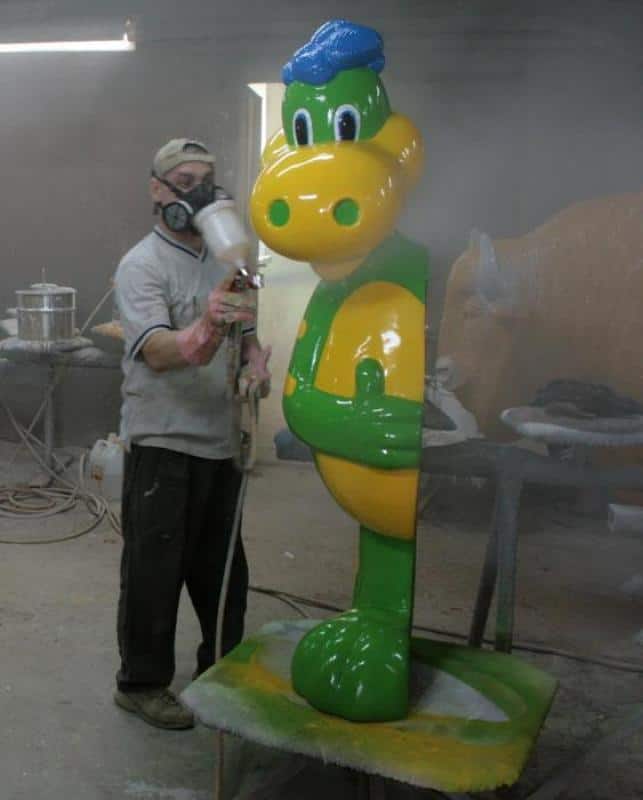 Mężczyzna maluje natryskowo dużą zielono-żółtą figurkę animowanej postaci w warsztacie.
