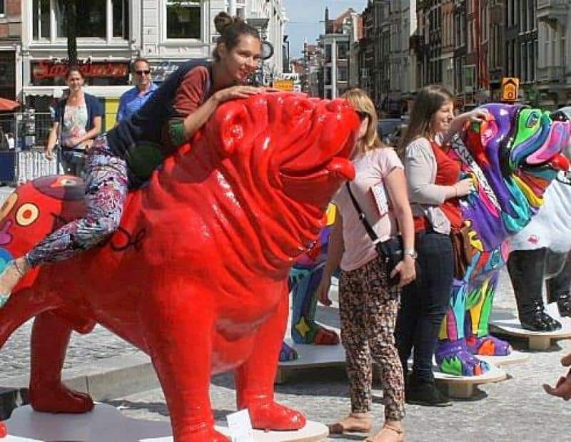 oryg_064490685_1129949550544944_1465385417682452480_o People interacting with large, colorful dog statues in a public square.